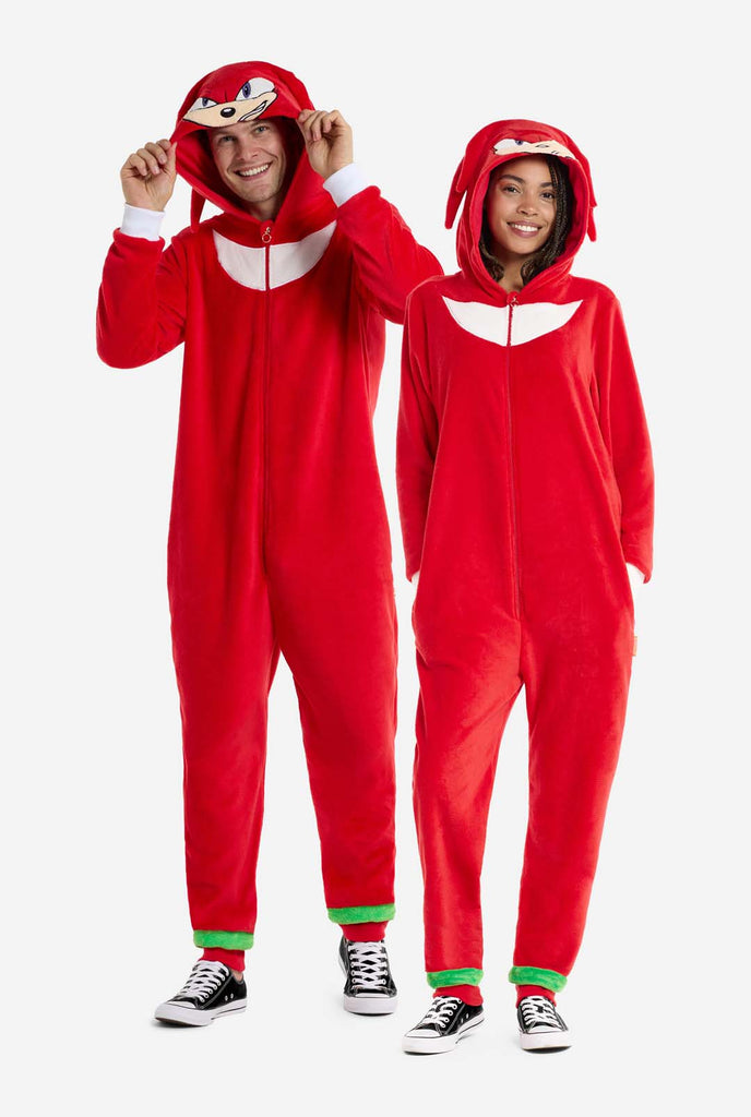 Een man en een vrouw dragen een felrode onesie voor volwassenen met Knuckles the Echidna-thema van OppoSuits. De leuke en knusse onesie heeft een ritssluiting aan de voorkant en een vaste capuchon die lijkt op het gezicht van Knuckles, compleet met expressieve ogen en slappe dreadlocks. Het pak is voornamelijk rood, met een wit halvemaanvormig patroon op de borst, wat de iconische look van Knuckles uit de videogameserie Sonic the Hedgehog weerspiegelt. Full body image.