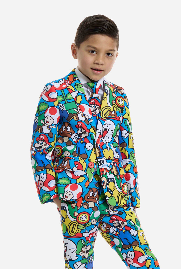 Afbeelding van kind dat een Super Mario all over print pak draagt, bestaande uit blazer, broek en stropdas. Hoofdkleur is blauw, maar er is ook geel, rood en wit.