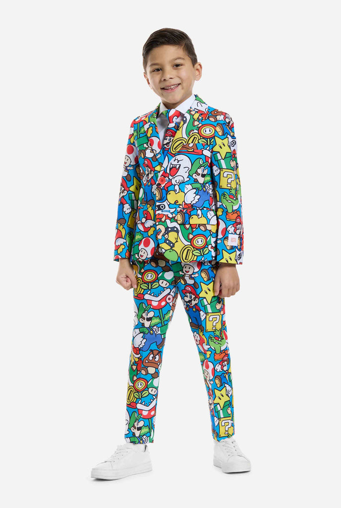 Afbeelding van kind dat een Super Mario all over print pak draagt, bestaande uit blazer, broek en stropdas. Hoofdkleur is blauw, maar er is ook geel, rood en wit.