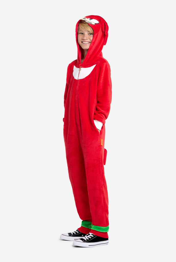 Een kind draagt ​​een felrode kinderonesie met Knuckles the Echidna-thema van OppoSuits. De leuke en knusse onesie heeft een ritssluiting aan de voorkant en een vaste capuchon die lijkt op het gezicht van Knuckles, compleet met expressieve ogen en slappe dreadlocks. Het pak is voornamelijk rood, met een wit halvemaanvormig patroon op de borst, wat de iconische look van Knuckles uit de videogameserie Sonic the Hedgehog weerspiegelt.