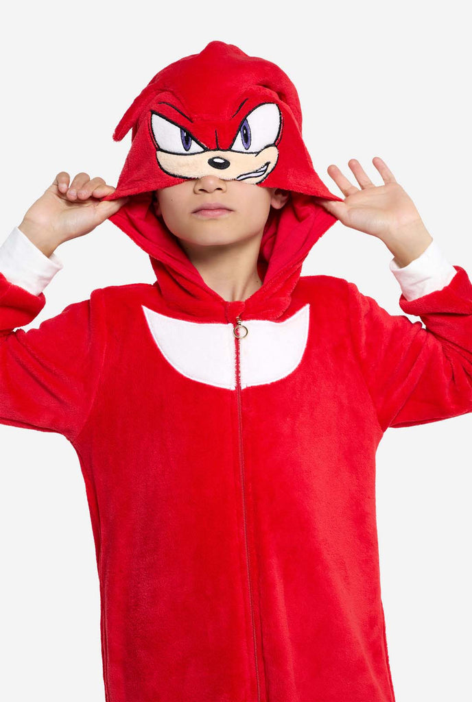 Een kind draagt ​​een felrode kinderonesie met Knuckles the Echidna-thema van OppoSuits. De leuke en knusse onesie heeft een ritssluiting aan de voorkant en een vaste capuchon die lijkt op het gezicht van Knuckles, compleet met expressieve ogen en slappe dreadlocks. Het pak is voornamelijk rood, met een wit halvemaanvormig patroon op de borst, wat de iconische look van Knuckles uit de videogameserie Sonic the Hedgehog weerspiegelt.