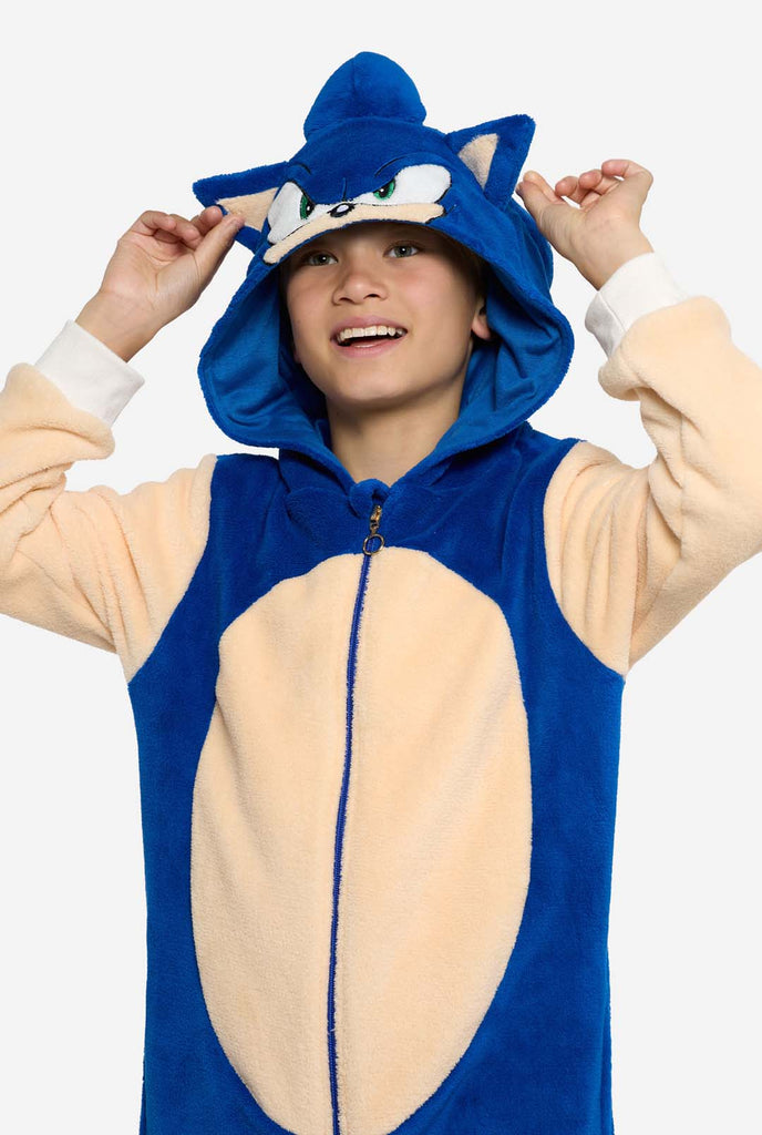 Een kind draagt ​​een blauw en beige Sonic the Hedgehog-thema kinderonesie van OppoSuits. De comfortabele onesie heeft een rits aan de voorkant en een capuchon die is ontworpen om te lijken op Sonics iconische gezicht, compleet met oren en een expressieve blik. Het blauwe lichaam van het pak contrasteert met de beige buik, wat Sonics look nabootst. Deze leuke, comfortabele en stijlvolle onesie is perfect voor loungen, themafeestjes en brengt nostalgische videogame-vibes naar elke gelegenheid.