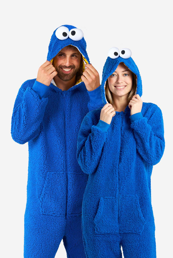 Man en vrouw dragen blauwe pluche Cookie Monster Adult onesie voor volwassenen. Staan naast elkaar, capuchon vasthoudend met de hand.