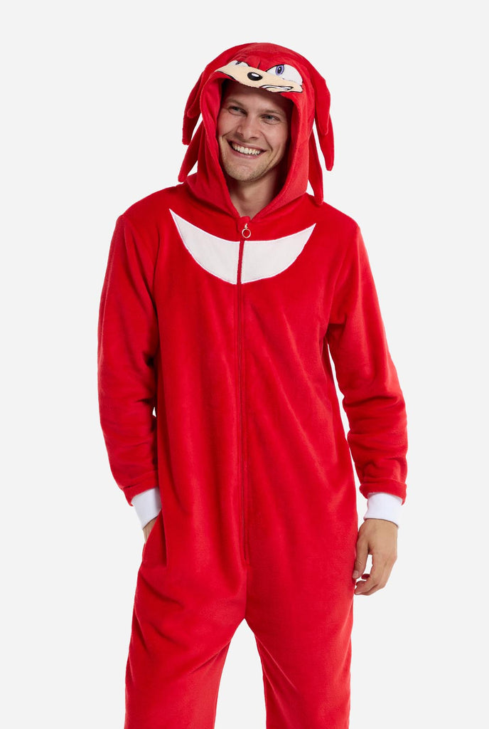 Een man draagt ​​een felrode onesie voor volwassenen met Knuckles the Echidna-thema van OppoSuits. De leuke en knusse onesie heeft een ritssluiting aan de voorkant en een vaste capuchon die lijkt op het gezicht van Knuckles, compleet met expressieve ogen en slappe dreadlocks. Het pak is voornamelijk rood, met een wit halvemaanvormig patroon op de borst, wat de iconische look van Knuckles uit de videogameserie Sonic the Hedgehog weerspiegelt.