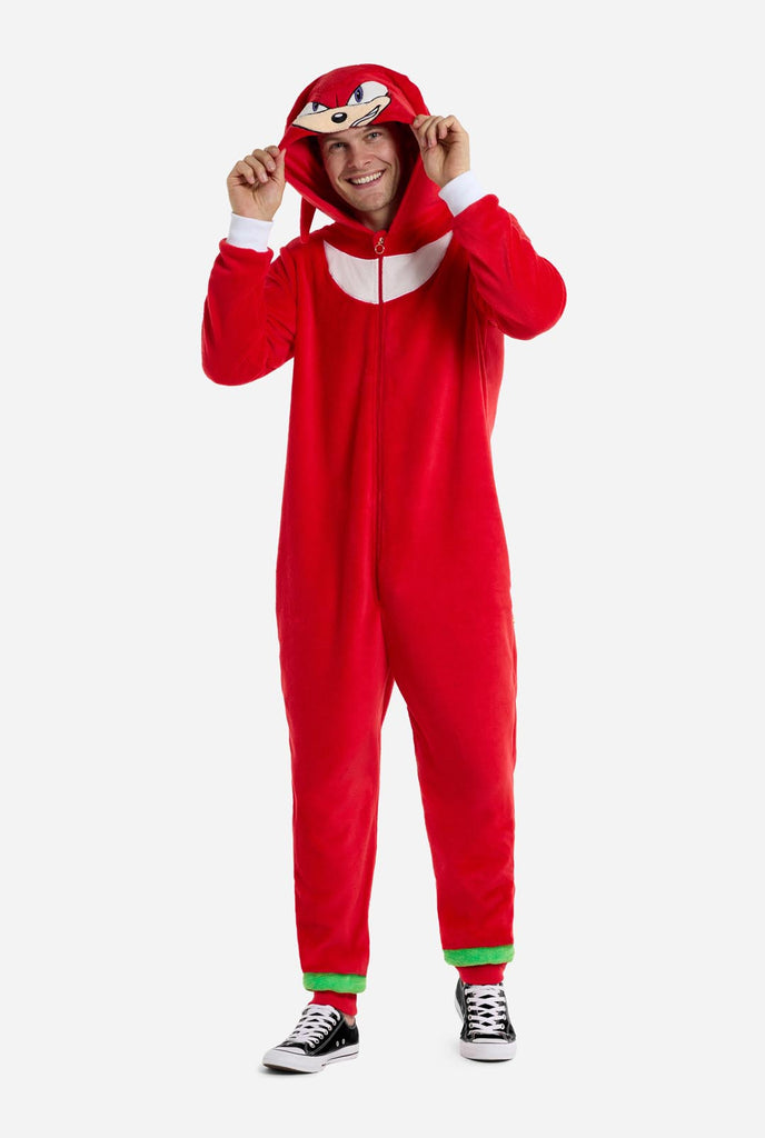 Een man draagt ​​een felrode onesie voor volwassenen met Knuckles the Echidna-thema van OppoSuits. De leuke en knusse onesie heeft een ritssluiting aan de voorkant en een vaste capuchon die lijkt op het gezicht van Knuckles, compleet met expressieve ogen en slappe dreadlocks. Het pak is voornamelijk rood, met een wit halvemaanvormig patroon op de borst, wat de iconische look van Knuckles uit de videogameserie Sonic the Hedgehog weerspiegelt.