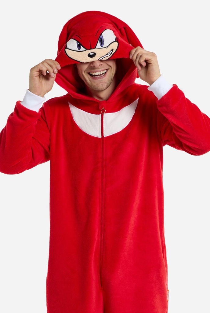 Een man draagt ​​een felrode onesie voor volwassenen met Knuckles the Echidna-thema van OppoSuits. De leuke en knusse onesie heeft een ritssluiting aan de voorkant en een vaste capuchon die lijkt op het gezicht van Knuckles, compleet met expressieve ogen en slappe dreadlocks. Het pak is voornamelijk rood, met een wit halvemaanvormig patroon op de borst, wat de iconische look van Knuckles uit de videogameserie Sonic the Hedgehog weerspiegelt.