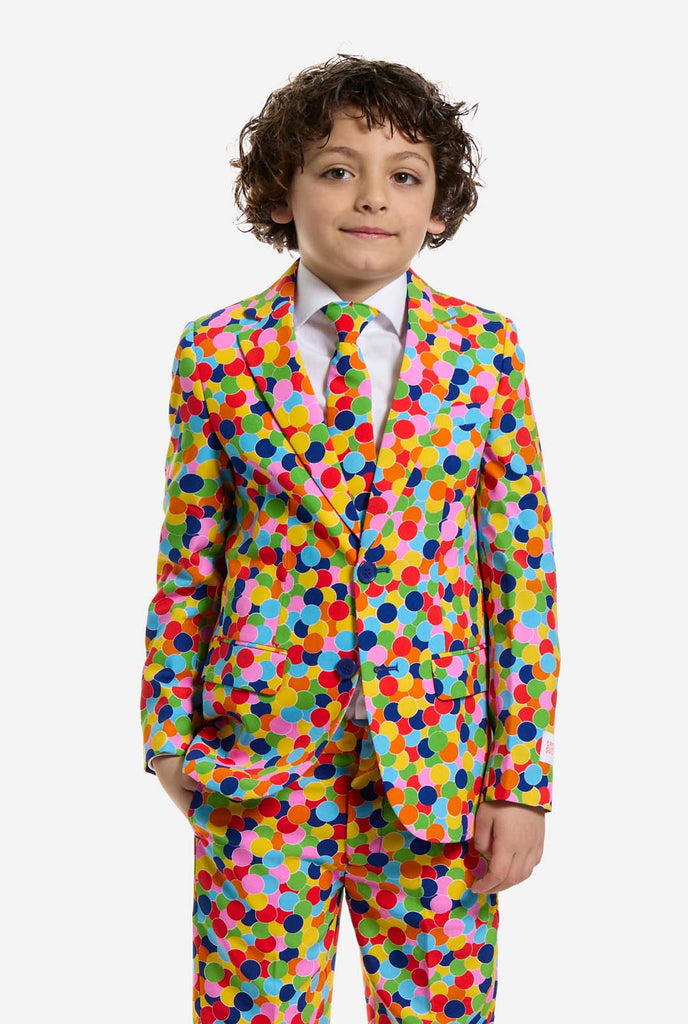 Op deze afbeelding draagt ​​een kind een jongenspak met all-over multicolor confetti print en een wit shirt. Het pak heeft een fel patroon met cirkels in verschillende kleuren, waaronder oranje, geel, roze, groen, blauw en rood.