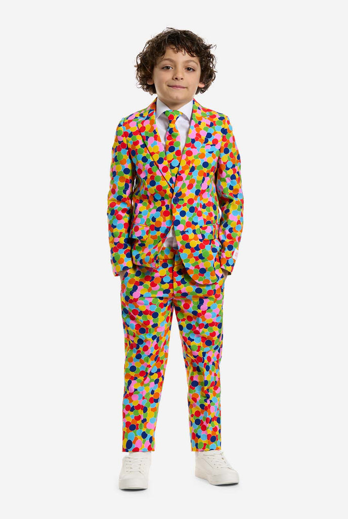 Op deze afbeelding draagt ​​een kind een jongenspak met all-over multicolor confetti print en een wit shirt. Het pak heeft een fel patroon met cirkels in verschillende kleuren, waaronder oranje, geel, roze, groen, blauw en rood.