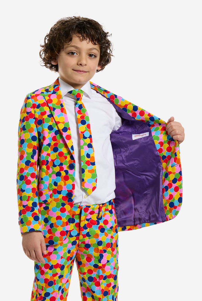 Op deze afbeelding draagt ​​een kind een jongenspak met all-over multicolor confetti print en een wit shirt. Het pak heeft een fel patroon met cirkels in verschillende kleuren, waaronder oranje, geel, roze, groen, blauw en rood. 1 kant van het jasje open.