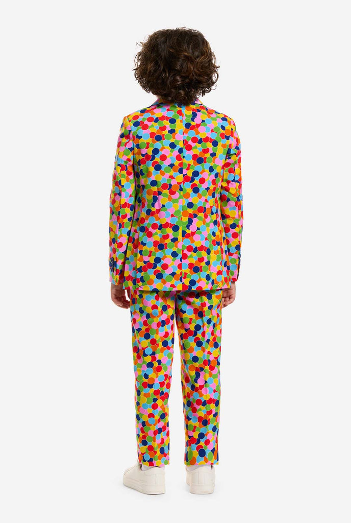 Op deze afbeelding draagt ​​een kind een jongenspak met all-over multicolor confetti print en een wit shirt. Het pak heeft een fel patroon met cirkels in verschillende kleuren, waaronder oranje, geel, roze, groen, blauw en rood. Beeld van de achterkant.