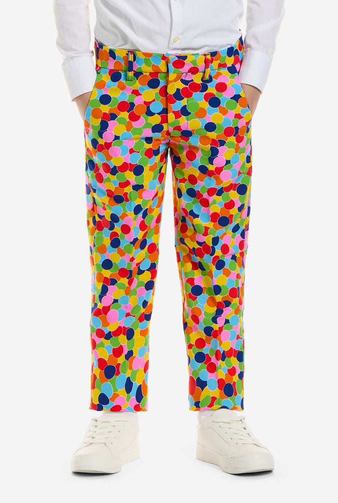 Op deze afbeelding draagt ​​een kind een jongenspak met all-over multicolor confetti print en een wit shirt. Het pak heeft een fel patroon met cirkels in verschillende kleuren, waaronder oranje, geel, roze, groen, blauw en rood. Close up broek.