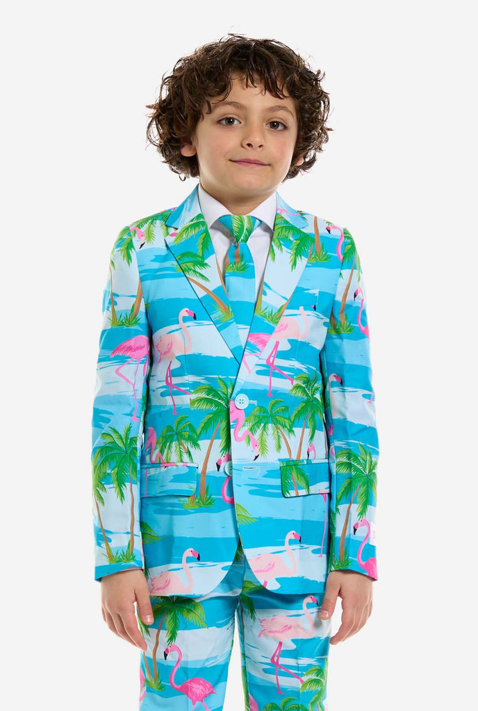 Op deze afbeelding draagt ​​een kind een lichtblauw jongenspak met een tropische print en een wit shirt. Het pak heeft een blauwe print die lijkt op water en roze flamingo's en groene palmbomen.