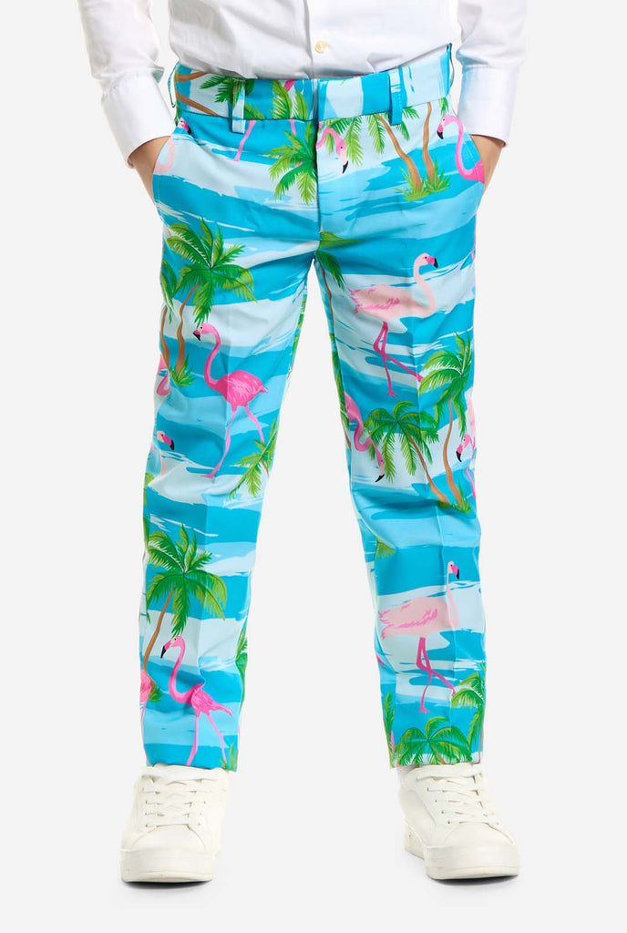 Op deze afbeelding draagt ​​een kind een lichtblauw jongenspak met een tropische print en een wit shirt. Het pak heeft een blauwe print die lijkt op water en roze flamingo's en groene palmbomen. Close up broek.