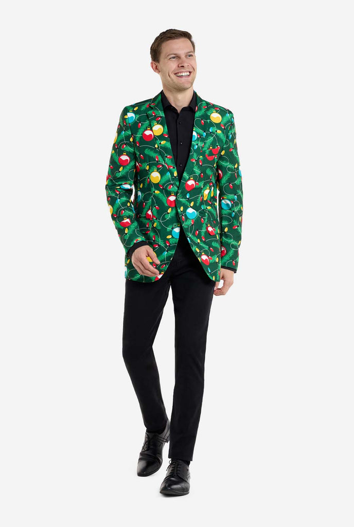 Man wearing green Christmas blazer for men with decorated Christmas tree print. Man also wearing a black pants and black men's shirt. Full body image.