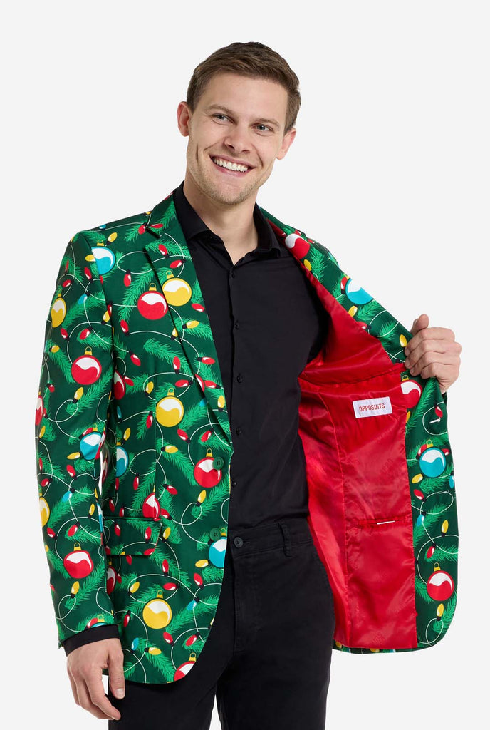 Man wearing green Christmas blazer for men with decorated Christmas tree print. Man also wearing a black pants and black men's shirt. Showing red inside of the jacket.