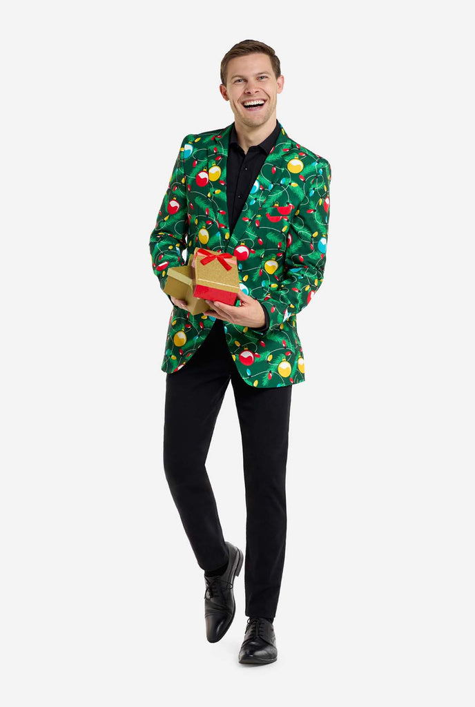 Man wearing green Christmas blazer for men with decorated Christmas tree print. Man also wearing a black pants and black men's shirt. Holding presents.