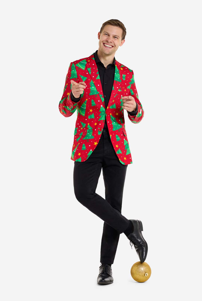 Man wearing red Christmas blazer for men with Christmas trees and stars print. Man also wearing a black pants and black men's shirt. Standing with one feet on Christmas ball. Full body image.