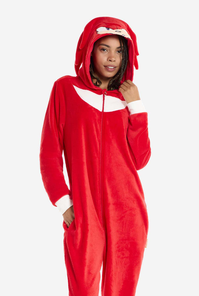 Een vrouw draagt ​​een felrode Knuckles the Echidna onesie voor volwassenen, een gezellige en speelse outfit geïnspireerd op het beroemde personage uit de Sonic the Hedgehog-serie. De onesie is voorzien van de kenmerkende witte borstpatch van Knuckles en heeft een capuchon die het gezicht van Knuckles laat zien, compleet met zijn expressieve ogen en lange rode dreads. De ritssluiting aan de voorkant zorgt voor gemakkelijk dragen, en de vrouw staat met één hand op haar zij.