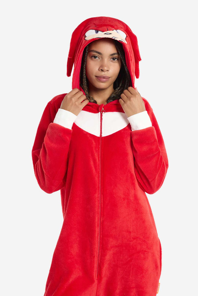 Een vrouw draagt ​​een felrode Knuckles the Echidna onesie voor volwassenen, een gezellige en speelse outfit geïnspireerd op het beroemde personage uit de Sonic the Hedgehog-serie. De onesie is voorzien van de kenmerkende witte borstpatch van Knuckles en heeft een capuchon die het gezicht van Knuckles laat zien, compleet met zijn expressieve ogen en lange rode dreads.