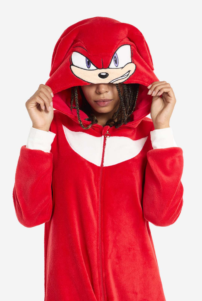 Een vrouw draagt ​​een felrode Knuckles the Echidna onesie voor volwassenen, een gezellige en speelse outfit geïnspireerd op het beroemde personage uit de Sonic the Hedgehog-serie. De onesie is voorzien van de kenmerkende witte borstpatch van Knuckles en heeft een capuchon die het gezicht van Knuckles laat zien, compleet met zijn expressieve ogen en lange rode dreads. De ritssluiting aan de voorkant zorgt voor een comfortabele pasvorm en de vrouw houdt de capuchon over haar ogen. 