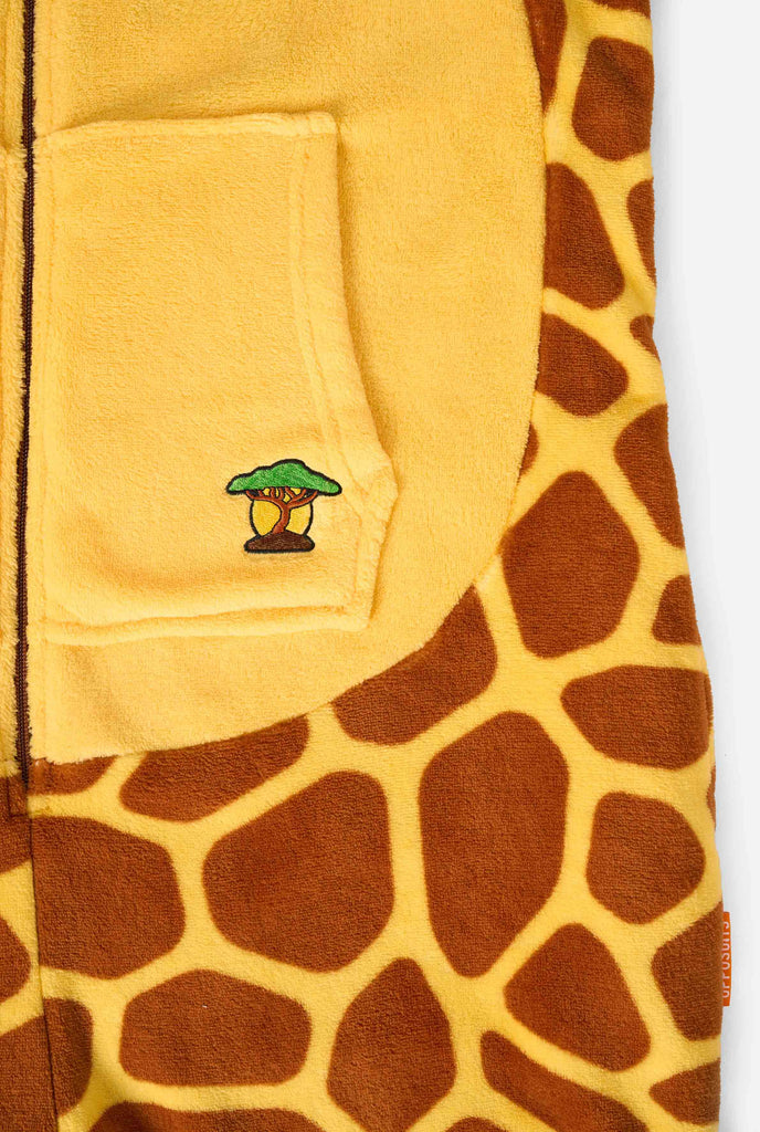 Een lachend kind draagt ​​een bruin en geel kinder onesie ontworpen als een giraffe. De onesie heeft een capuchon die lijkt op het hoofd van een giraffe.